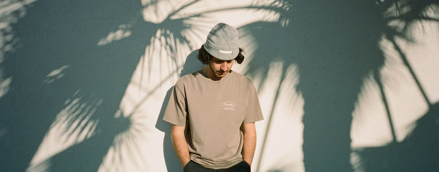Model in a grey Simple beanie and a sand oval t-shirt against a wall with palm tree shadows
